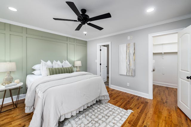 bedroom with a white bed green accent wall nightstands with lamps a ceiling fan and wooden flooring there is an open door leading to a walk in closet and a door to another room