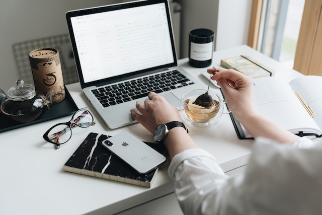 person sitting at a desk working on a macbook dunking a tea bag with an open notebook glasses and phone on the desk